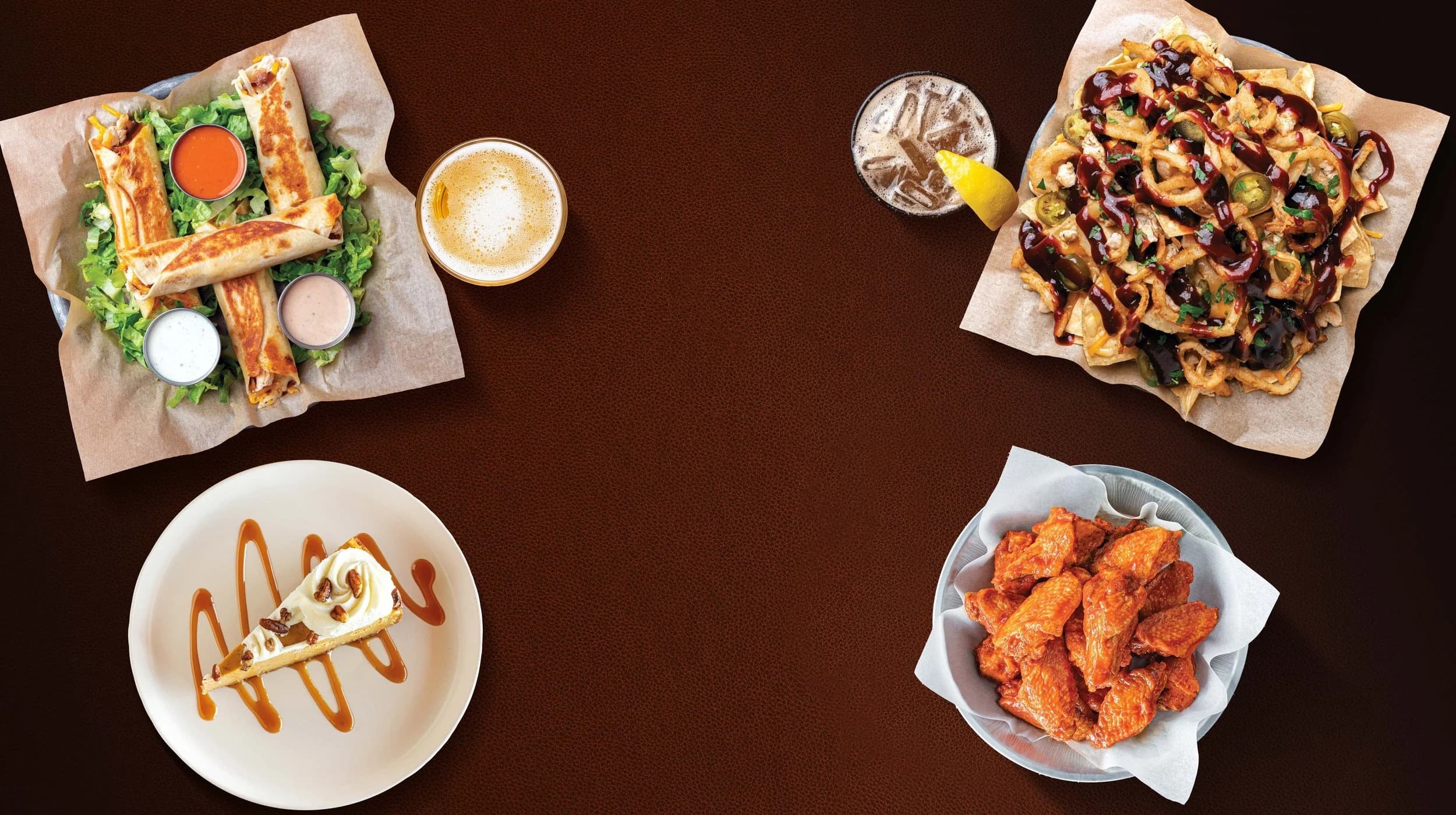 Game-day spread on a dark tabletop: chicken wraps, loaded nachos, wings, cheesecake, a beer and an iced tea